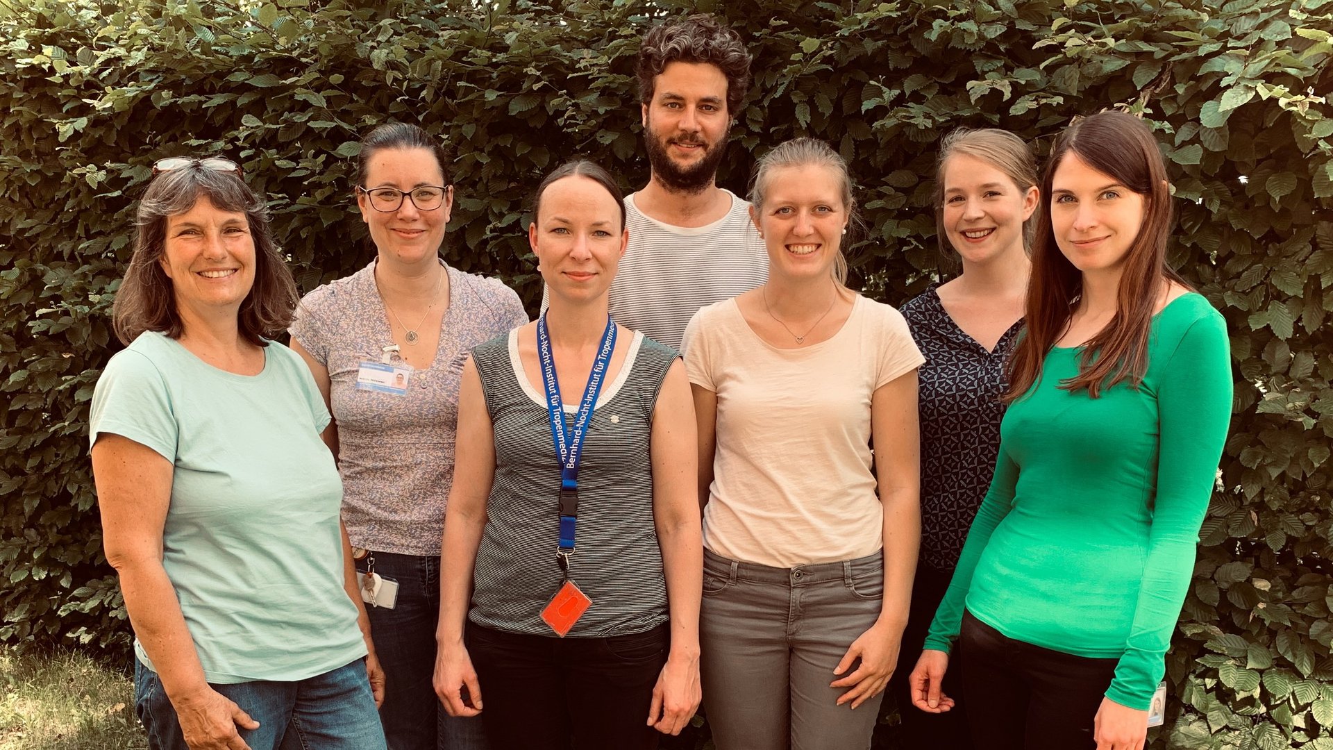 Fröhliches frontales Gruppenfoto der Gruppe um Maria Rosenthal vor einer grnen Hecke.