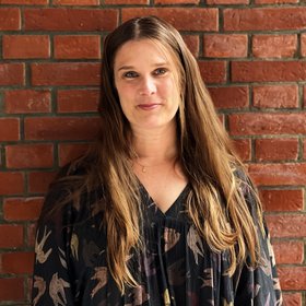 Anna Bachmann: a researcher stands outside in front of a brick wall and laughs into the camera.