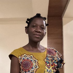 Das Bild zeigt eine Frau mit kurzen schwarzen Haaren, die freundlich in die Kamera blickt. Sie trägt ein gelbes Shirt mit rotem Muster.