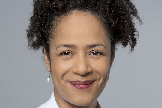 Prof. Dr. Marylyn Addo Prof. Dr. Marylyn Addo: Das Bild zeigt eine Ärztin in weißem Arztkittel mit lockigen schwarzen Haaren, die zu einem Zopf zurückgebunden sind und weißen Ohrringen.
