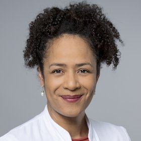 Prof. Marylyn Addo: The picture shows a female doctor in a white doctor's coat with curly black hair tied back in a braid and white earrings.