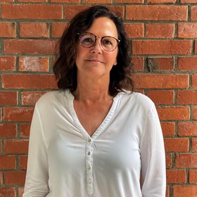 Heidrun von Thien: a technical assistant with medium-length hair and brown-rimmed glasses in front of a brick wall.