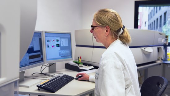 An technical assistant sits at a computer and looks at the screen. A large gray device can be seen behind her.