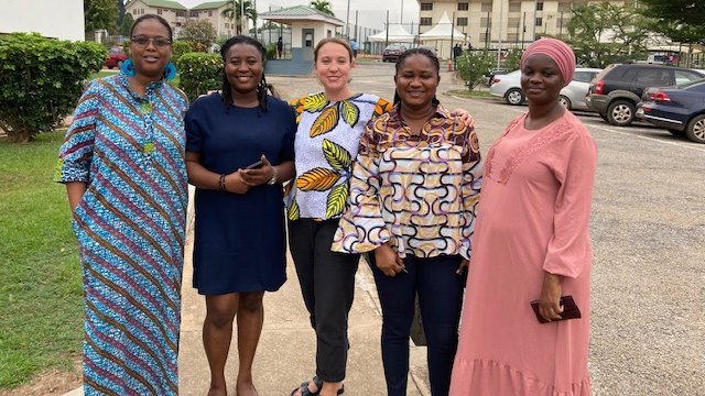 Kontrollbesuch in Ghana: eine diverse Gruppe von fünf Frauen steht auf einem Gehweg neben einer Straße und lachen in die Kamera. Im Hintergrund stehen Häuser mit grüne Vorgärten, auf der Straße parken Autos und der Himmel ist bewölkt.