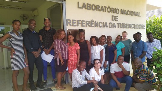 Staff of the LNR-TB in São Tomé  Das Foto zeigt sechzehn Sao Tome und Principe Mitarbeiter:innen des Laboratório Nacional de Referência da Tuberculose zusammen mit zwei Mitarbeitern des BNITM vor dem Laborgebäude in São Tomé und Príncipe. Die Gruppe steht vor einer Hasuwand, auf der der Schriftzug "Laboratório Nacional de Referência da Tuberculose" zu sehen ist.