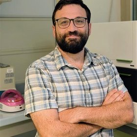 A man with glasses and a beard sitting at a desk, smiling with arms crossed. He is wearing a plaid shirt. In the background, laboratory equipment is visible, including a device with a pink lid.