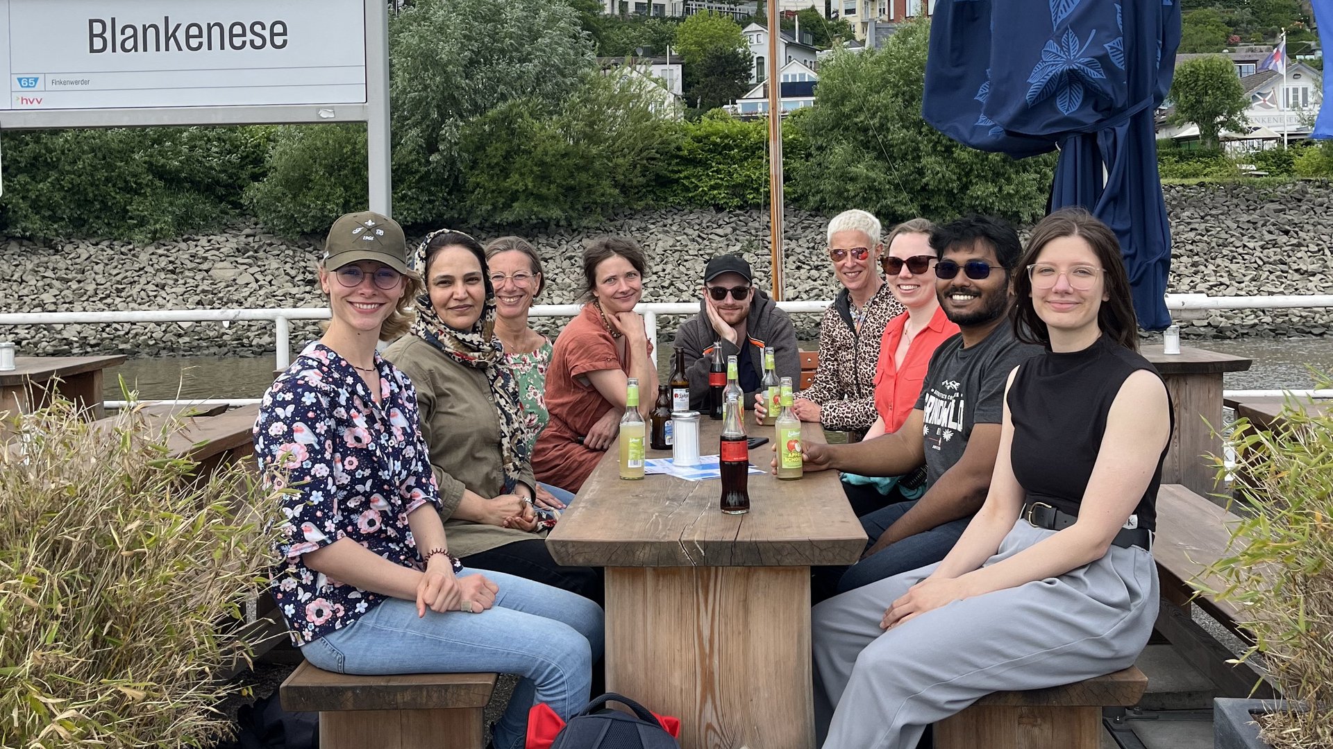 A group of smiling people sitting around a table outdoors next to a sign saying "Blankenese"