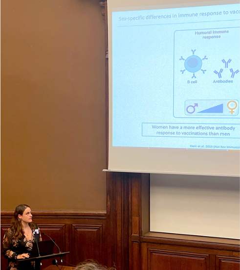 The picture shows Charlotte Hansen in a lecture hall at the Pasteur Institute. She is small in the bottom left of the picture, while her presentation is large in the background. Charlotte is wearing a dark dress with flowers.