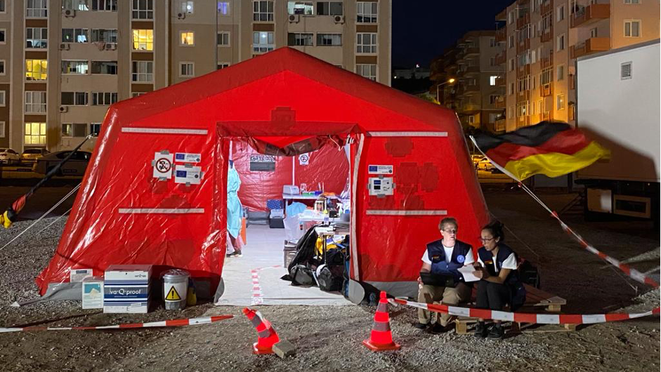 The image is shot in the dark. In the foregound is a large red tent with a brightly lit lab inside. Behind the tent blocks of flats are visible.