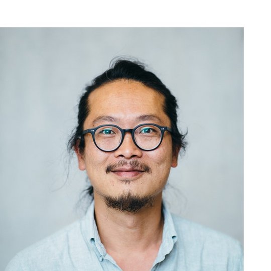 Portrait of a male person of color with glasses and dark hair pulled back, wearing a blue shirt.