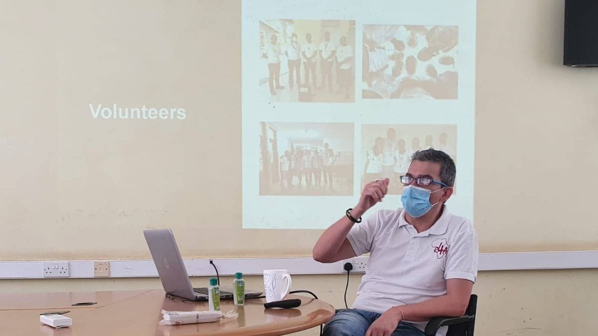 Training and capacity building during the hospital partnership workshop in Tanzania Zu sehen ist ein Mann, der vor einem an die Wand gebeamten Bild sitzend einen Vortrag hält. Man erkennt das Wort Volunteers auf der Projektion. Er trägt einen Nasen-Mund-Schutz.