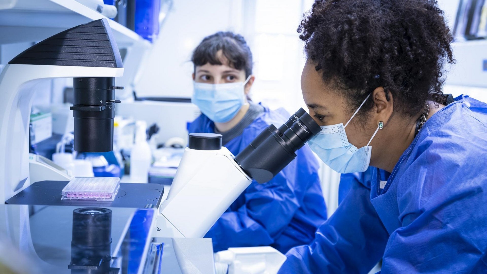 Two researchers in a laboratory wearing blue protective clothing. The researcher in the foreground is looking into a microscope.