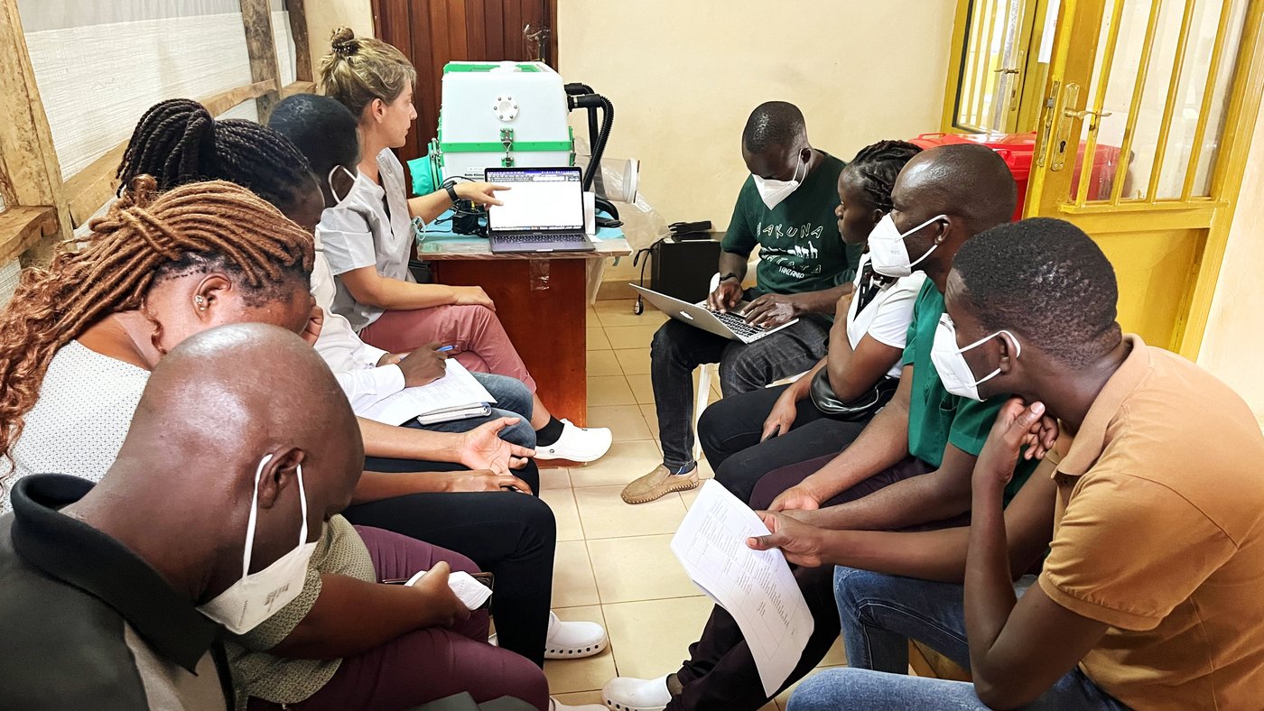 A room with nine laboratory team members sitting in a circle discussing the implementation of new standard operating procedures (SOP) in the CPHL-EAC mobile laboratory during the Sudan Ebola virus outbreak in Uganda. One person leads the discussion using a computer on which agenda items are displayed. The other staff members take notes or listen (Mubende, November 2022; CPHL: Central Public Health Laboratories, EAC: East Africa Community).