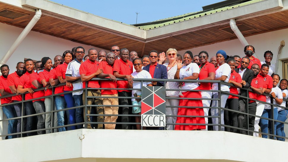 Mitarbeitende des KCCR in überwiegend roten T-Shirts stehen auf dem Balkon des Instituts, einige winken. Vorn in der Mitte stehen der wissenschaftliche Direktor Prof. Phillips und die blonde Geschäftsführerin Ingrid Sobel.