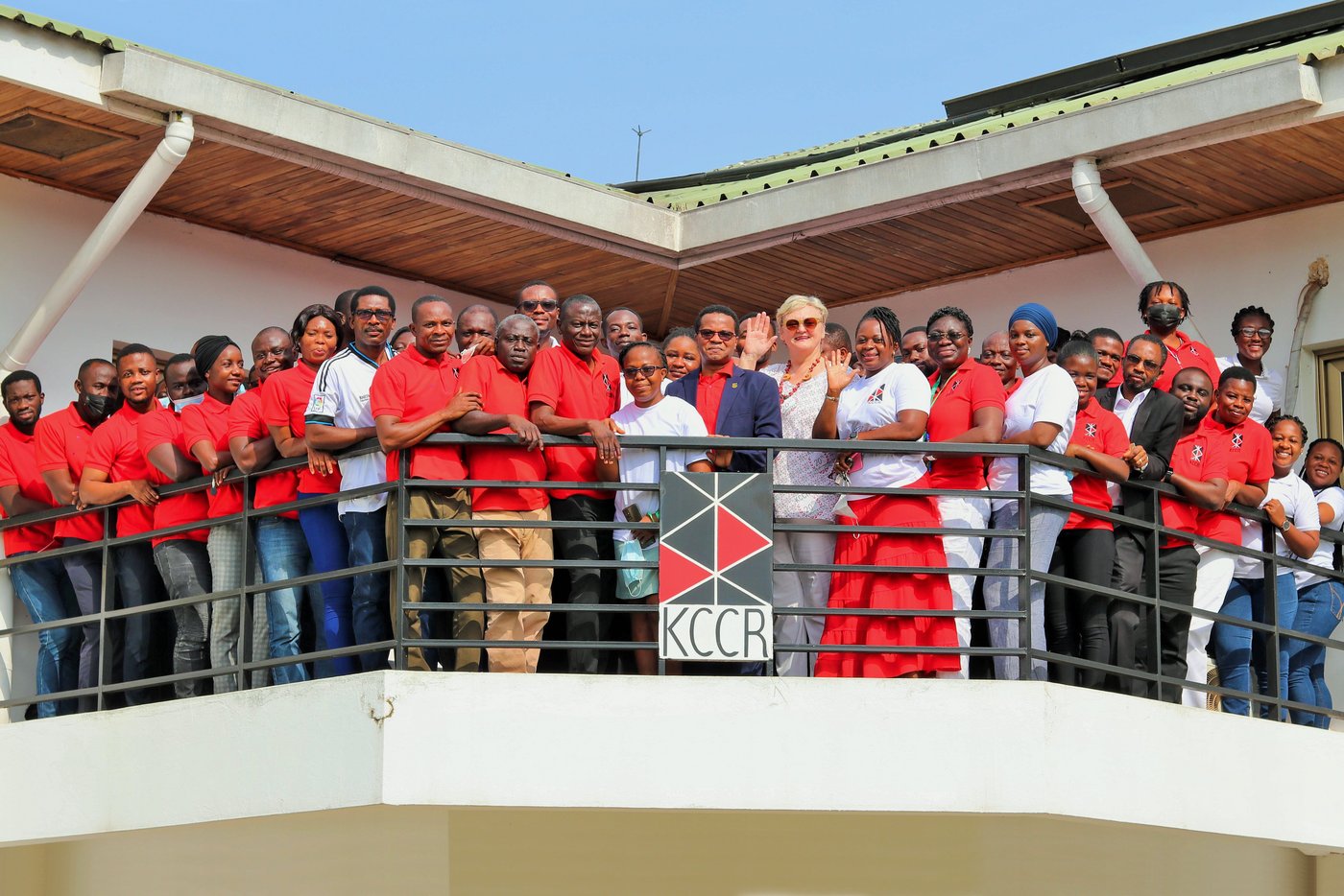 Das Team des KCCR Mitarbeitende des KCCR in überwiegend roten T-Shirts stehen auf dem Balkon des Instituts, einige winken. Vorn in der Mitte stehen der wissenschaftliche Direktor Prof. Phillips und die blonde Geschäftsführerin Ingrid Sobel.