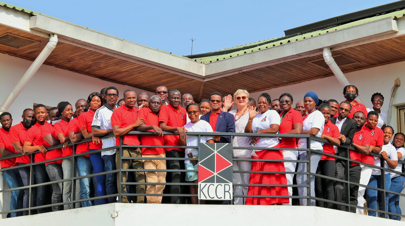 Mitarbeitende des KCCR in überwiegend roten T-Shirts stehen auf dem Balkon des Instituts, einige winken. Vorn in der Mitte stehen der wissenschaftliche Direktor Prof. Phillips und die blonde Geschäftsführerin Ingrid Sobel.
