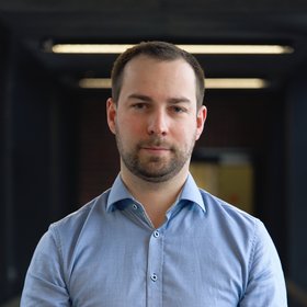 Direct photo of Till Omansen, you see a researcher in a light blue shirt against a dark background