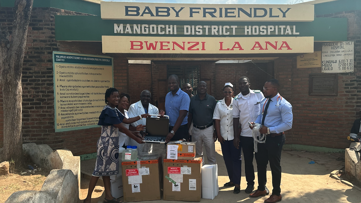 The picture shows nine people standing in front of a hospital. In front of them are packages and an ultrasound machine is visible which is being held by two people.