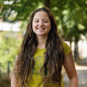 Sarah Ryter: eine lächelnde Frau mit langen Haaren in einem grünen T-Shirt auf einem sonnigen Weg unter Bäumen.