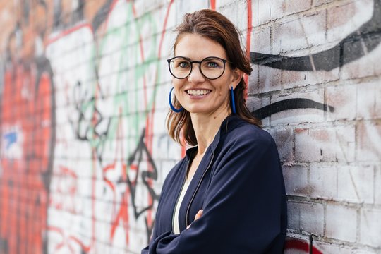 Prof. Dr Cornelia Betsch A photo of Cornelia Betsch in front of a graffiti-covered brick wall. She is wearing a dark blue suit, has shoulder-length dark brown hair and distinctive dark-framed glasses, and is smiling warmly at the camera.
