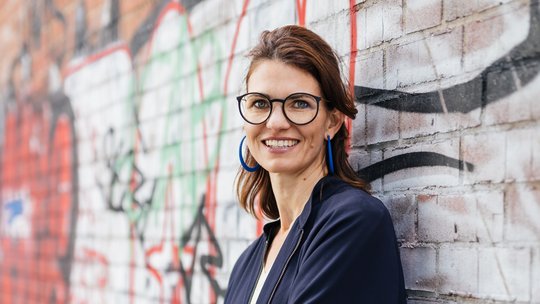Prof. Dr. Cornelia Betsch Foto von Cornelia Betsch vor einer Graffiti-Backsteinwand. Sie trägt ein dunkelblaues Kostüm, schulterlange dunkelbraune Haare, eine markante dunkel umrahmte Brille und blickt freundlich in die Kamera.