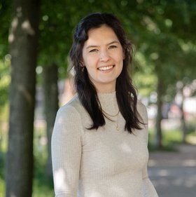Jasmin Scharnberg Photo of a smiling young woman with long, dark hair wearing a beige pullover. She is standing in the shade of green trees. fy,
