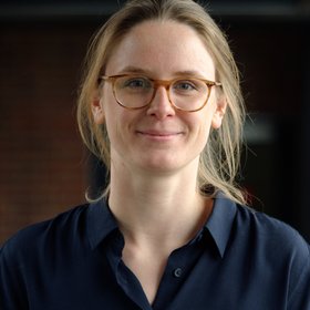A women in her end twenties infront of a black background. The woman wears glasses and has blond hair.