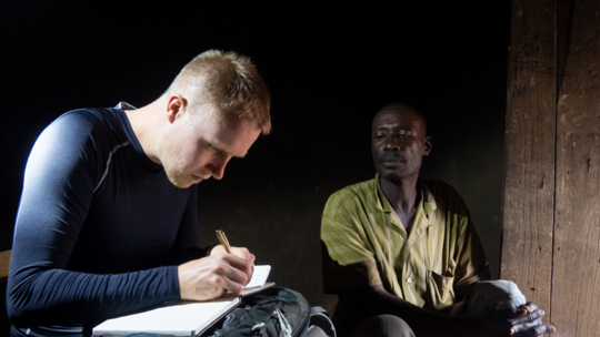Fabien Schultz ist links im Vordergrund zu sehen, wie er sich Notizen macht. Rechts im Hintergrund sitzt ein traditioneller Heiler.