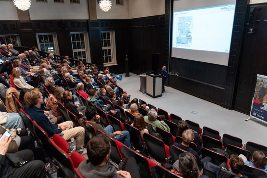 Das Foto zeigt den fast voll besetzten Hörsaal des BNITM. Vorne steht eine Rednerin am Pult, im Hintergrund sieht man eine Folie der Präsentation.
