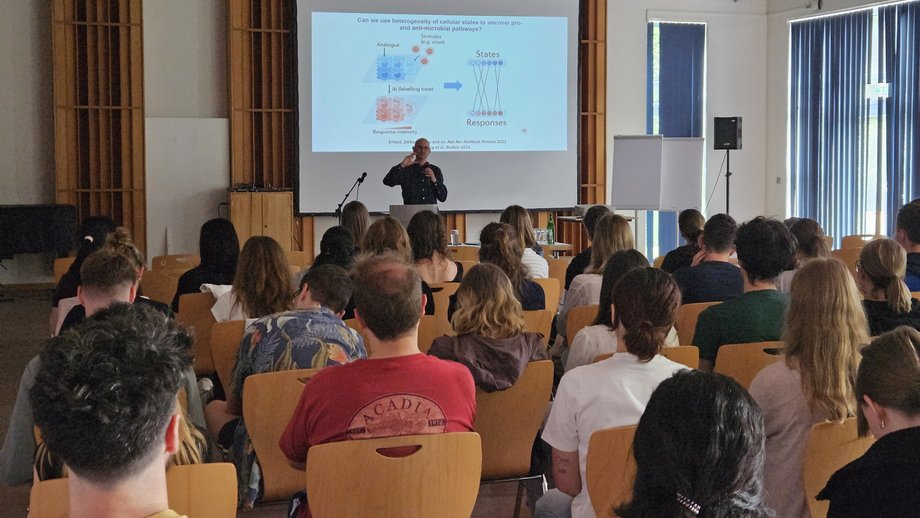 Blick von hinten auf Studierende, die einem Vortrag in einem hellen Seminarraum mit Holzdecke und großen Fenstern folgen. Eine Person steht vorne und hält einen Vortrag, auf einer Leinwand dahinter ist eine Folie mit biologischem Schaubild zu sehen.