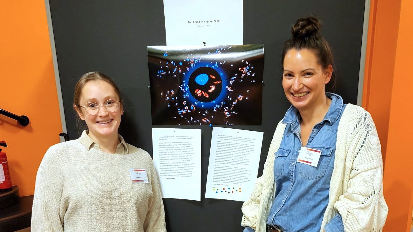 Zwei Forscherinnen stehen vor einer Aufstellwand, auf der ein Bild mit schwarzem Hintergrund zu erkennen, auf dem unterschiedlich geformte in blau und rot gehaltene Zellen gezeigt sind.