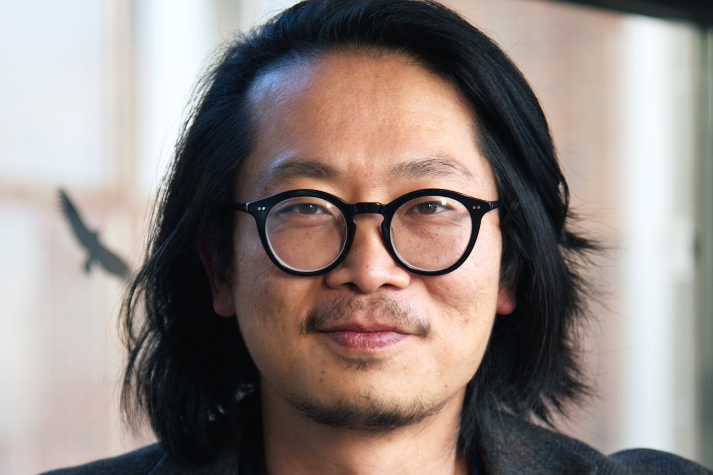 Dr. Sung-Joon Park: a researcher who wears shoulder-length hair, round black glasses, a dark jacket over a dark blue top.