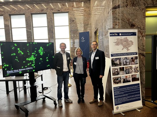 Das Foto zeigt drei Personen zwischen einem Monitor mit Zellbild und einem Forschungsposter stehen.