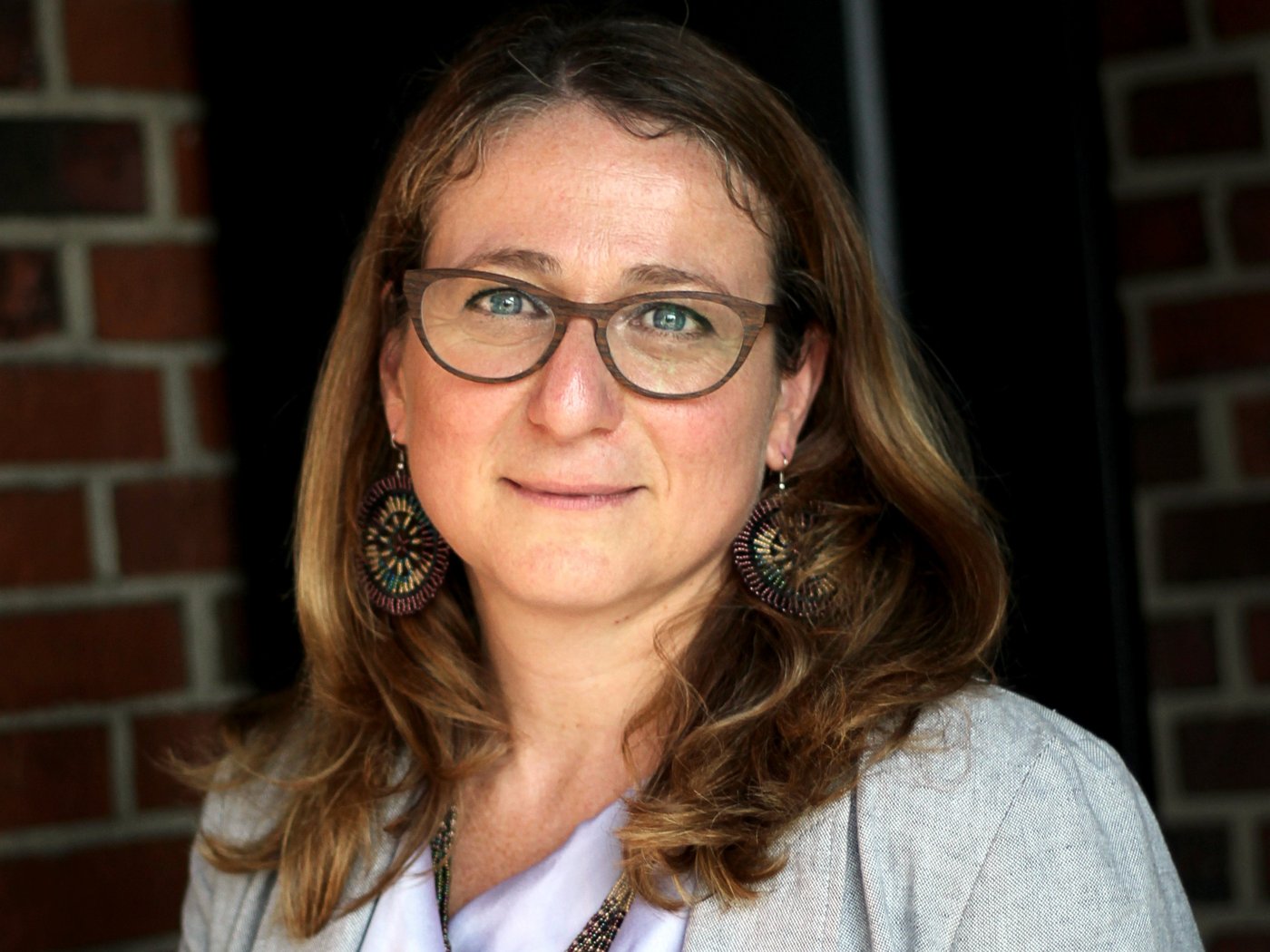 Daniela Fusco, Ph.D. Daniela Fusco, Ph.D.: a researcher against a dark red brick background, wearing her blonde shoulder-length hair loose.