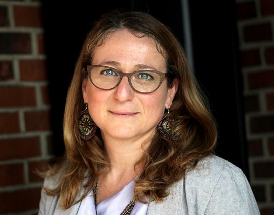 Daniela Fusco, Ph.D.: eine Forscherin vor dunklen roten Backstein Hintergrund, die ihre blonden schulterlangen Haar offen trägt.