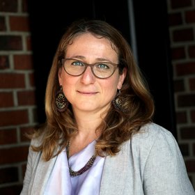 Daniela Fusco, Ph.D. Daniela Fusco, Ph.D.: a researcher against a dark red brick background, wearing her blonde shoulder-length hair loose.