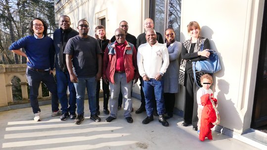 Das Foto zeigt eine Gruppe diverser Forschender. Sie stehen draußen auf einer Terrasse in der Sonne und lächeln.