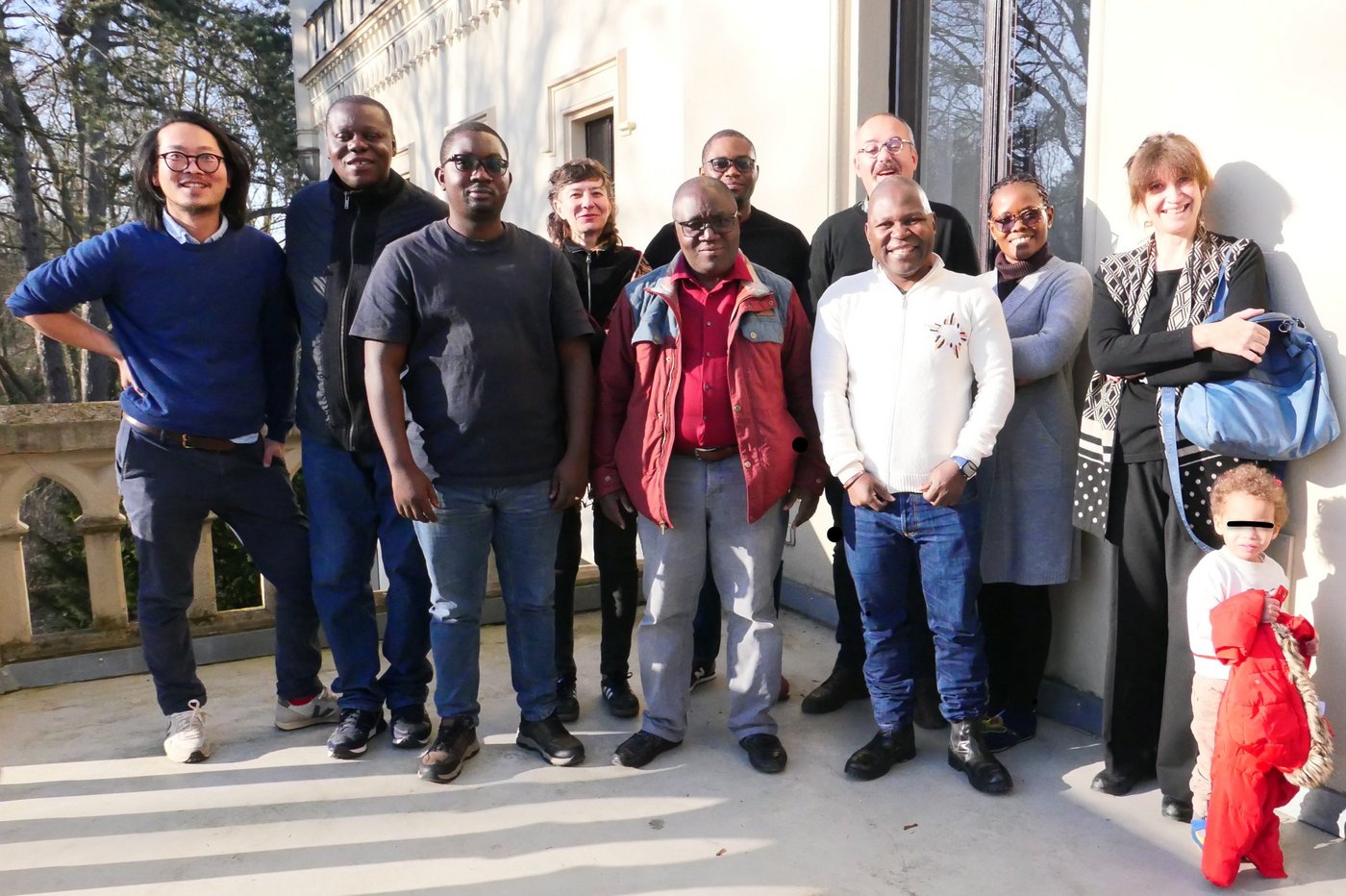 The photo shows a group of diverse researchers. They are standing outside on a terrace in the sun and smiling.