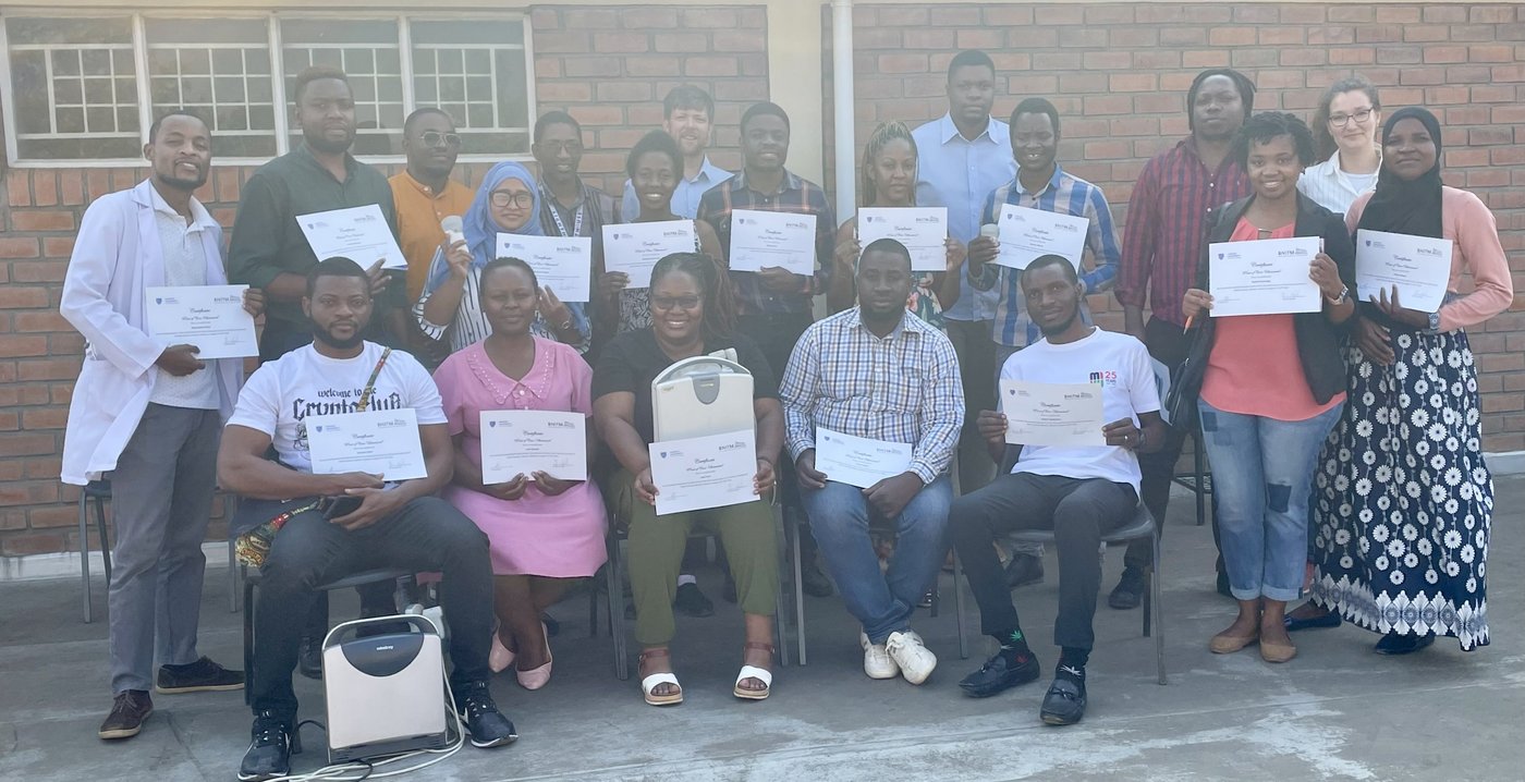 The picture shows a group of twenty people standing and sitting in front of a brick building. Some people are holding certificates and ultrasound machines.