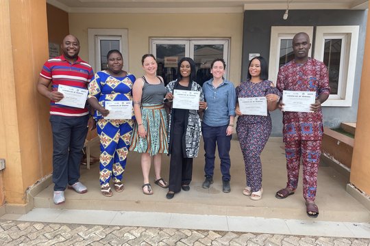 Sequencing lab staff from ISTH together with BNITM trainers at the end of the 2-weeks metagenomics sequencing training at ISTH. February 2024, Irrua, Nigeria. Five Nigerian laboratory staff and two BNITM trainers in colourful clothes are standing in the entrance of a building. They are proudly holding up their training certificates.
