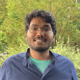 Saleh Yunus: A PhD student smiles at the camera. He wears medium length black hair, glasses, a short beard and a light blue casual shirt.