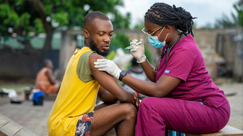 Foto einer jungen afrikanischen Krankenschwester mit Gesichtsmaske, die einem jungen Schwarzen draußen eine Spritze gibt.