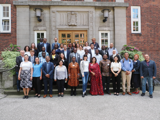 Alumni group photo Gruppenfoto von etwa 40 Personen vor einem historischen Backsteingebäude mit Holztür und Steinfassade. Die vielfältige Gruppe umfasst Männer und Frauen unterschiedlichen Alters und ethnischer Herkunft, die in Geschäfts- oder halbformeller Kleidung gekleidet sind und freundlich in die Kamera lächeln. Die Szene deutet auf eine akademische oder berufliche Veranstaltung hin.