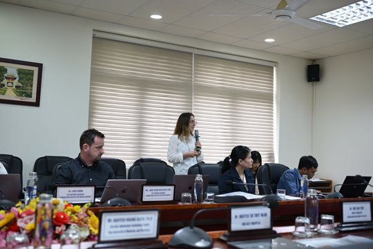 The picture shows a woman speaking into the microphone while three other people look at the presentation.