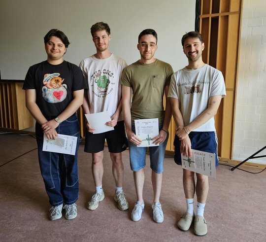 Preise LCI Summer School 2026 Auf dem Foto stehen vier junge Männer, die Urkunden in den Händen halten. Alle haben kurze Haare, tragen T-Shirts sowie kurze Hosen, bis auf eine Person mit langer Hose.