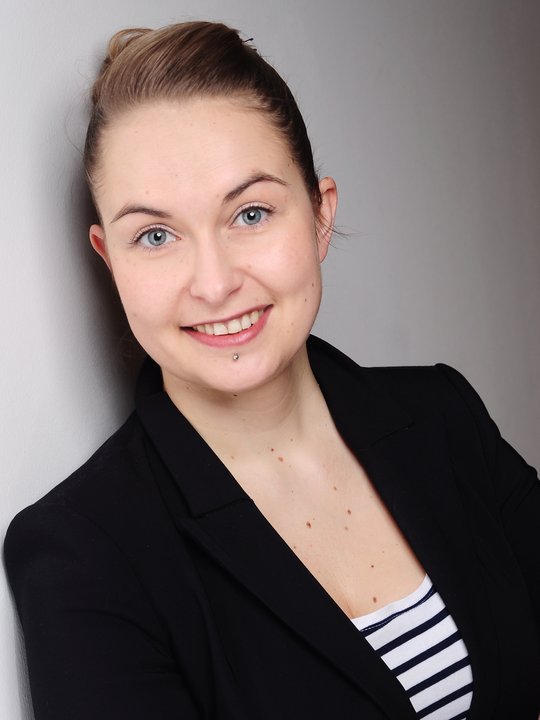 Dr. Anna Heitmann Dr. Anna Heitmann: Eine Forscherin mit dunkelblonden Haaren, das zu einem Zopf gebunden ist. Sie trägt einen schwarzen Blazer, darunter ein blau weiß gestreiftes Oberteil und ist an eine graue Betonwand gelehnt.