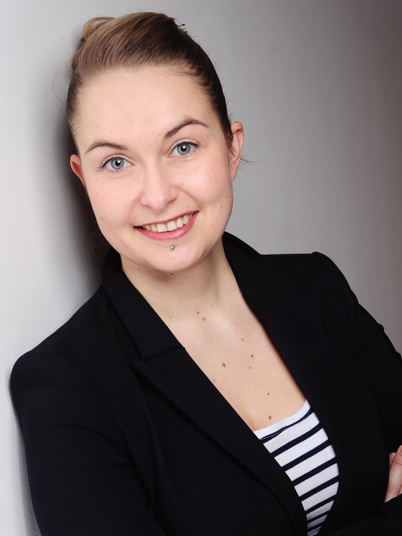 Dr Anna Heitmann Dr Anna Heitmann: A researcher with dark blonde hair tied back in a ponytail. She is wearing a black blazer over a blue and white striped top and is leaning against a grey concrete wall.
