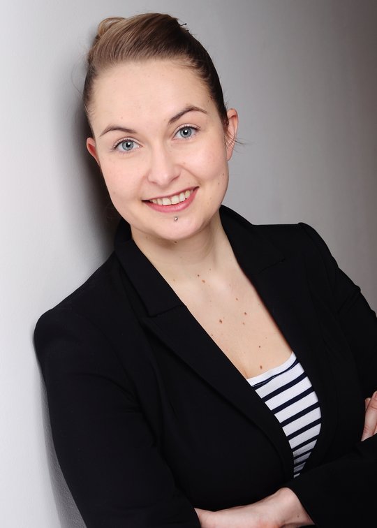 Dr. Anna Heitmann: Eine Forscherin mit dunkelblonden Haaren, das zu einem Zopf gebunden ist. Sie trägt einen schwarzen Blazer, darunter ein blau weiß gestreiftes Oberteil und ist an eine graue Betonwand gelehnt.