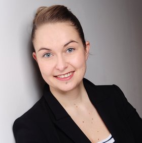 Dr Anna Heitmann: A researcher with dark blond hair tied in a braid, wearing a black blazer, a blue and white striped top underneath and leaning against a white wall.