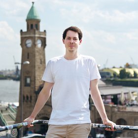 Maximilian Guigas: a PhD student wearing longer brown hair and a light-coloured top stands in front of the ancient building of the Landungsbrücken.